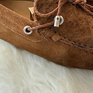 May include: Close-up of a pair of brown suede loafers. The shoes feature a braided leather lace detail with silver-toned metal eyelets and accents. The suede has a textured appearance.