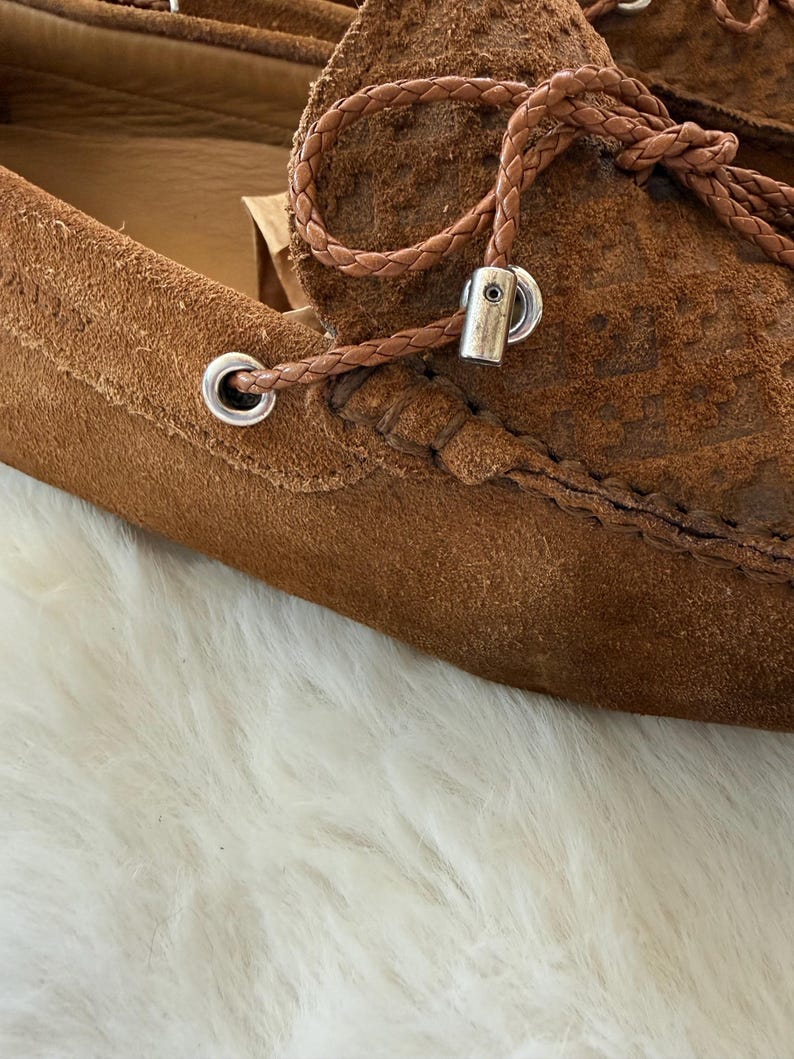 May include: Close-up of a pair of brown suede loafers. The shoes feature a braided leather lace detail with silver-toned metal eyelets and accents. The suede has a textured appearance.
