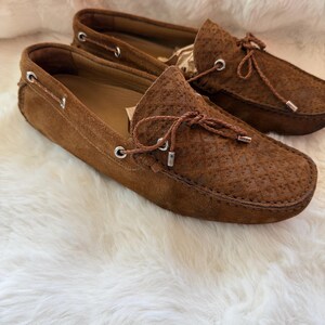 May include: A pair of brown suede loafers with decorative laces and silver-toned eyelets. The shoes have a textured pattern on the upper and are set against a white, fluffy background. The classic design suggests a focus on comfort and style.