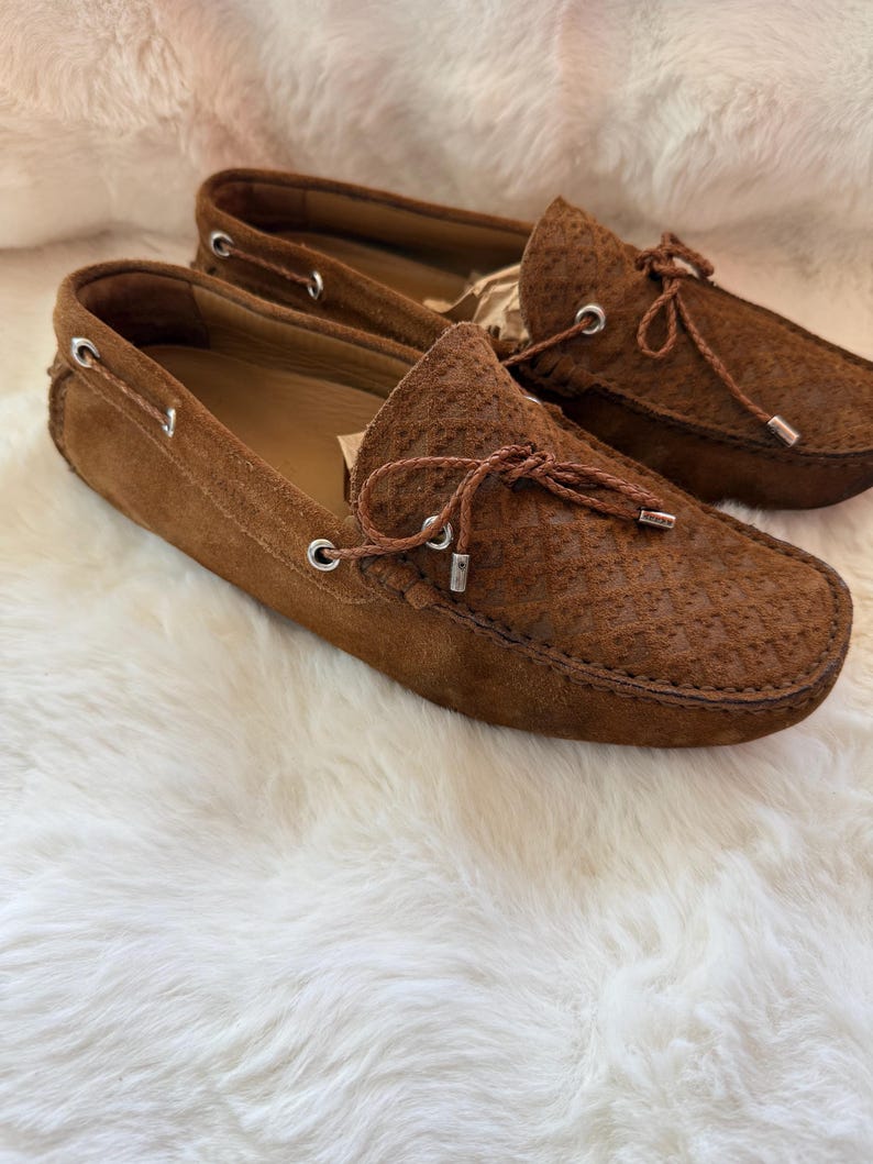 May include: A pair of brown suede loafers with decorative laces and silver-toned eyelets. The shoes have a textured pattern on the upper and are set against a white, fluffy background. The classic design suggests a focus on comfort and style.