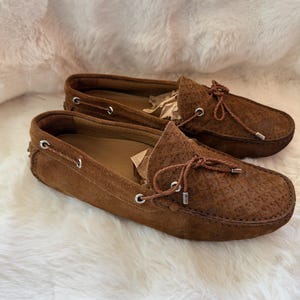 May include: A pair of brown suede loafers with a textured pattern on the toe box. The shoes have a leather tie detail and silver eyelets. The loafers are displayed on a white, fluffy surface.