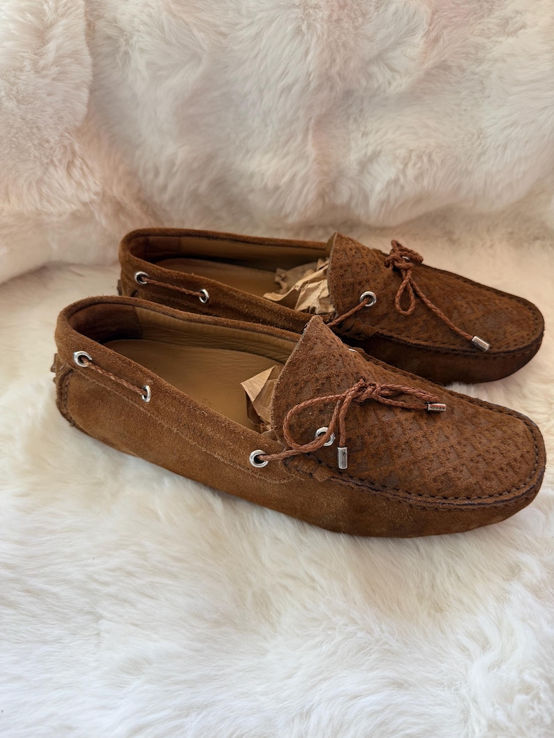 May include: A pair of brown suede loafers with a textured pattern on the toe box. The shoes have a leather tie detail and silver eyelets. The loafers are displayed on a white, fluffy surface.