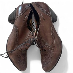 May include: Brown leather ankle boots with a lace-up design and a block heel. The boots feature decorative stitching and perforations along the edges. The brand name "Tamaris" is visible on the side.