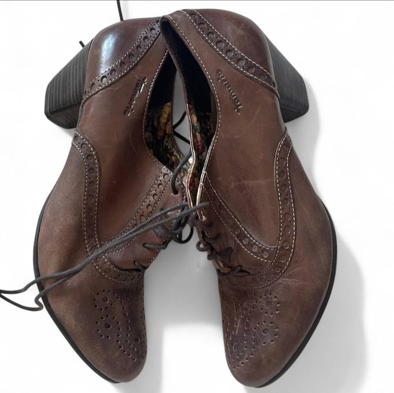 May include: Brown leather ankle boots with a lace-up design and a block heel. The boots feature decorative stitching and perforations along the edges. The brand name "Tamaris" is visible on the side.