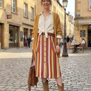 K&ouml;nnte beinhalten: Eine Frau tr&auml;gt eine senfgelbe Strickjacke, ein cremefarbenes Hemd und einen bunten gestreiften Rock. Sie tr&auml;gt auch braune Cowboy-Stiefel und eine braune Tragetasche mit Fransen.