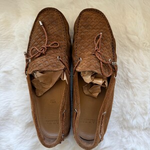 May include: A pair of brown suede loafers with a textured pattern. The shoes have leather laces and metal eyelets. The interior of the shoes is a lighter brown color. The shoes are displayed on a white, fluffy surface.
