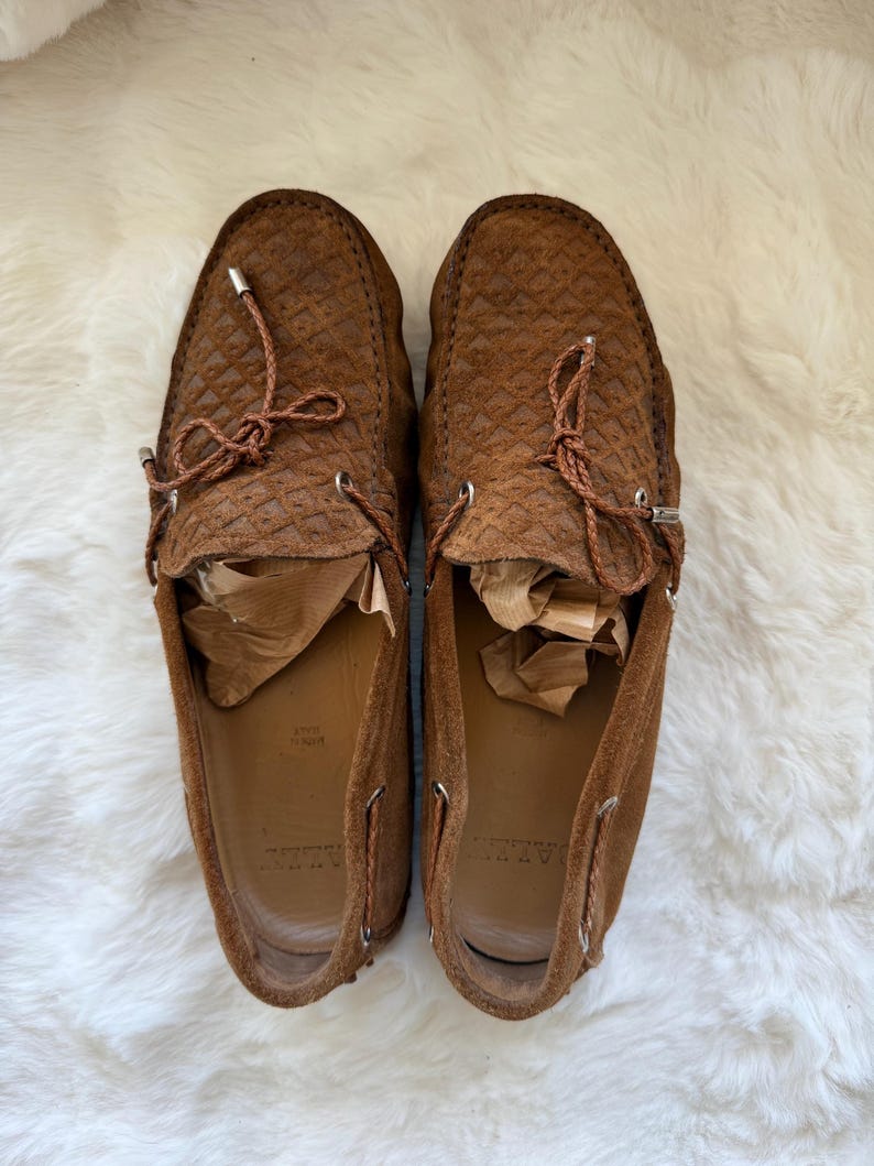 May include: A pair of brown suede loafers with a textured pattern. The shoes have leather laces and metal eyelets. The interior of the shoes is a lighter brown color. The shoes are displayed on a white, fluffy surface.