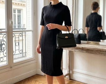 May include: A black knee-length dress with a high neckline and short sleeves, paired with a structured black handbag featuring a gold-tone clasp and black pointed-toe high heels. The ensemble is completed with brown-tinted cat-eye sunglasses.