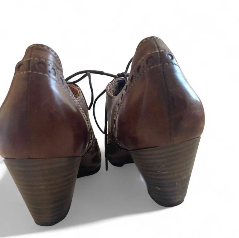May include: A pair of brown leather lace-up shoes with stacked heels. The shoes have decorative stitching and eyelets along the top edge. The heels are approximately 6.35 cm high.