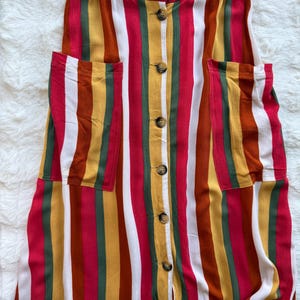 K&ouml;nnte beinhalten: Ein bunt gestreiftes Kleid mit Knopfleiste und zwei Vordertaschen. Das Kleid hat vertikale Streifen in Rot, Wei&szlig;, Gelb, Gr&uuml;n und Braun. Das Kleid hat einen gelben Bund und ist aus leichtem Stoff gefertigt.