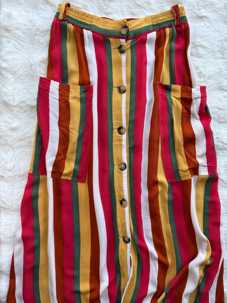 K&ouml;nnte beinhalten: Ein bunt gestreiftes Kleid mit Knopfleiste und zwei Vordertaschen. Das Kleid hat vertikale Streifen in Rot, Wei&szlig;, Gelb, Gr&uuml;n und Braun. Das Kleid hat einen gelben Bund und ist aus leichtem Stoff gefertigt.