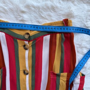 K&ouml;nnte beinhalten: Nahaufnahme eines farbenfrohen gestreiften Kleidungsst&uuml;cks mit Knopfleiste. Die Streifen umfassen Rot, Wei&szlig;, Gelb und Gr&uuml;n. Ein blaues Ma&szlig;band zeigt eine Messung von ca. 76 cm. Das Kleidungsst&uuml;ck hat zwei Taschen.