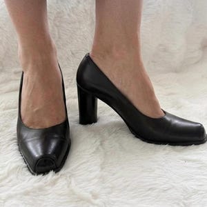 May include: A pair of black leather pumps featuring a square toe design and a sturdy block heel. The shoes have a smooth finish and a structured silhouette, providing a classic look with a distinctively shaped toe box and a solid, thick heel.