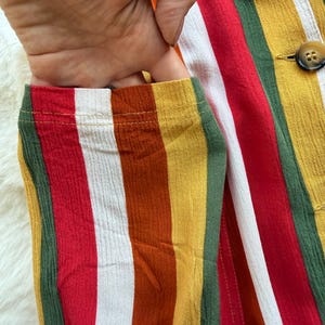 K&ouml;nnte beinhalten: Nahaufnahme eines farbenfrohen gestreiften Kleidungsst&uuml;cks mit einer Tasche. Der Stoff weist vertikale Streifen in Rot, Wei&szlig;, Gelb, Gr&uuml;n und Orange auf. Eine Hand ist teilweise sichtbar und greift in die Tasche. Zwei braune Kn&ouml;pfe sind sichtbar.