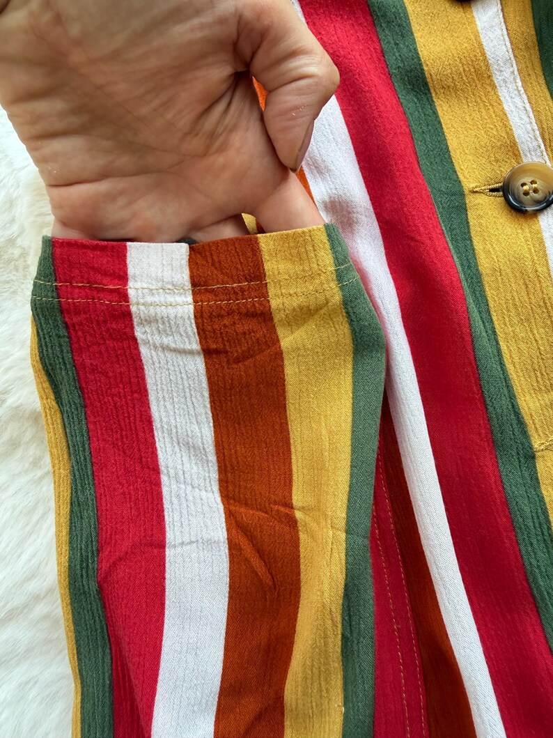 K&ouml;nnte beinhalten: Nahaufnahme eines farbenfrohen gestreiften Kleidungsst&uuml;cks mit einer Tasche. Der Stoff weist vertikale Streifen in Rot, Wei&szlig;, Gelb, Gr&uuml;n und Orange auf. Eine Hand ist teilweise sichtbar und greift in die Tasche. Zwei braune Kn&ouml;pfe sind sichtbar.