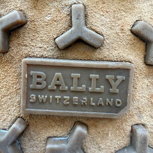 May include: Close-up of a shoe sole with a textured, light brown suede-like surface. The sole features raised, gray, Y-shaped treads and a rectangular, embossed label that reads "BALLY SWITZERLAND" in capital letters.