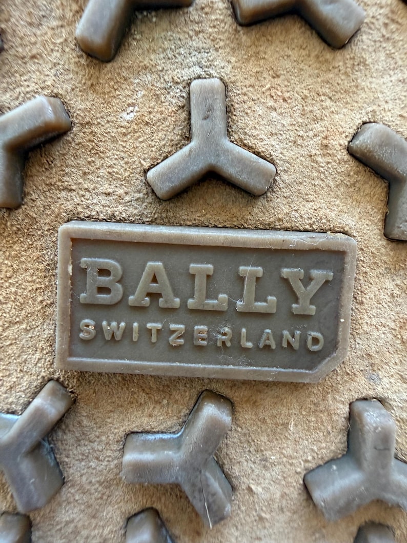 May include: Close-up of a shoe sole with a textured, light brown suede-like surface. The sole features raised, gray, Y-shaped treads and a rectangular, embossed label that reads "BALLY SWITZERLAND" in capital letters.