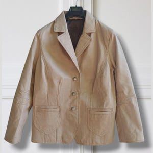 May include: A tan leather blazer with a notched collar, three buttons, and two front pockets. The jacket is hanging on a black hanger against a white background. The blazer has a classic, tailored design.