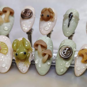 May include: Ten miniature nail art designs featuring various nature-inspired themes. The designs include mushrooms, a frog, a button, a dragonfly, and a flower.