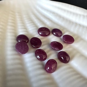 Può includere: Una collezione di gemme ovali, rosso scuro, disposte in un piatto bianco a forma di conchiglia. Le gemme hanno un aspetto lucido e brillante, che riflette la luce. Il piatto ha una superficie strutturata.