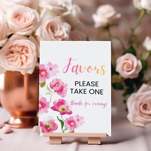 May include: A white sign with the word "Favors" in pink and yellow script, with the text "PLEASE TAKE ONE" and "thanks for coming!" in black. Pink watercolor flowers decorate the sign, which is displayed on a small wooden easel.