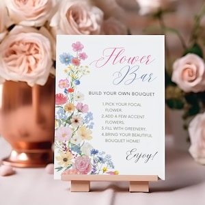 May include: A white sign with the words "Flower Bar" in pink and blue script, with instructions to build a bouquet. The sign is on a small wooden easel, with a copper vase of roses in the background.
