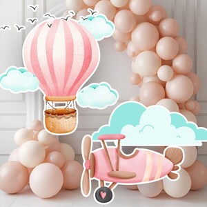 以下が含まれることがあります： ピンク色の熱気球、ピンク色の飛行機、ふわふわの雲が描かれた気まぐれなイラスト。 バルーンはストライプのデザインと織りバスケットです。 飛行機にはプロペラと車輪にハートが付いています。 ソフトピンクと白の風船がアーチを形成しています。