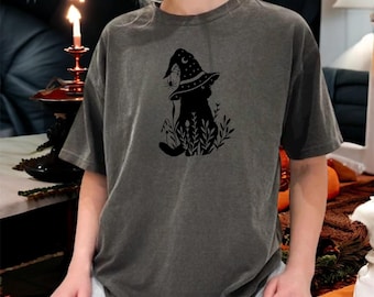 May include: Dark grey t-shirt featuring a black graphic of a cat wearing a witch hat with a spider and moon design. The cat is surrounded by foliage. The shirt has short sleeves and a relaxed fit.