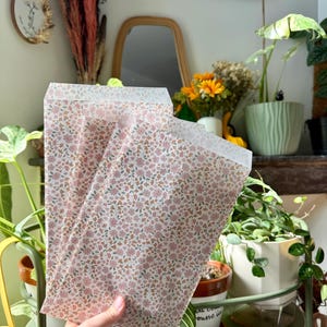 Peut inclure: Un ensemble de trois sacs en papier rectangulaires arborant un motif répétitif de petites fleurs roses et orange avec des feuilles vertes sur fond blanc. Les sacs sont semi-translucides et présentent une ouverture plate à bord droit.