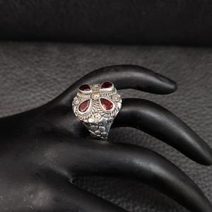 May include: A silver ring with a floral design, featuring four red gemstones and a gold accent in the center. The ring has an ornate, textured band. The ring is displayed on a black hand.