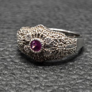 May include: A silver ring with an ornate filigree design. The ring features a central, round, deep pink gemstone, surrounded by smaller clear stones. The band has a detailed, textured pattern.