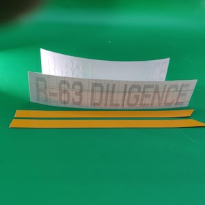 May include: Two white paper strips with the text "R-63 DILIGENCE" printed on them. Two yellow adhesive strips are also included.