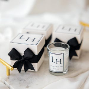 May include: White gift boxes tied with black ribbon bows and a small glass candle. Each box and candle features the initials "LH" in a black font. The candle also has the names "Juana & Hermano" and the date "11-02-2025".