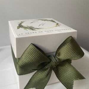 Puede incluir: Una caja de regalo blanca atada con un lazo de cinta verde. La tapa de la caja presenta un diseño de corona con marco dorado con el texto "10 YEARS OF GODS GOODNESS". La caja es de forma cuadrada y el lazo está hecho de tela acanalada.