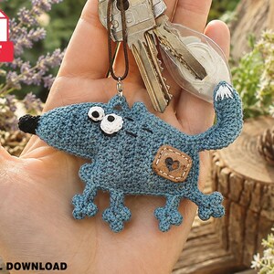 May include: A blue crocheted wolf keychain featuring a black nose, white eyes with black pupils, and a small brown square patch with a heart design on its side. The wolf has four small legs and a curved tail, attached to a metal keyring with a brown cord.