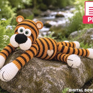 May include: A crochet tiger toy featuring orange and black stripes, a white muzzle, a black nose, and green eyes. The tiger has white paws and a small beige heart patch on its side. This item is a digital download PDF pattern for a stuffed animal.