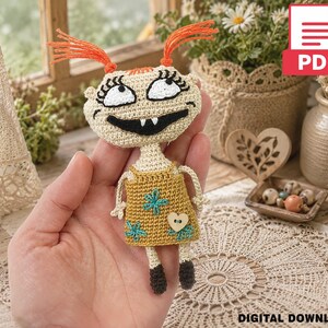 May include: A crochet doll featuring a beige head with orange yarn pigtails, large white eyes with black pupils, and a wide black mouth with two teeth. The doll wears a mustard yellow dress decorated with blue flower embroidery and a small heart button.