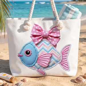 May include: White canvas tote bag with rope handles, featuring a whimsical fish applique with a pink polka dot bow. The fish has blue, pink, and white chevron patterned scales. A beach towel is visible inside the bag, and sunscreen and seashells are in the sand.