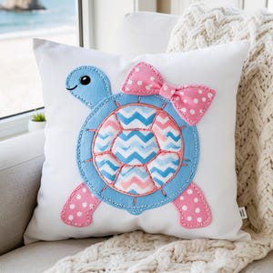 May include: A white square throw pillow featuring a central appliqué of a light blue turtle. The turtle shell has a blue and pink chevron pattern, while the head and limbs are accented with pink polka-dot fabric and a pink polka-dot bow.