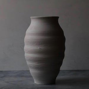 May include: A matte light gray ceramic vase with a rounded, bulbous body and a narrow neck. The surface features horizontal, undulating ridges and a textured finish created by visible wheel-thrown marks, giving the vessel a tactile appearance.
