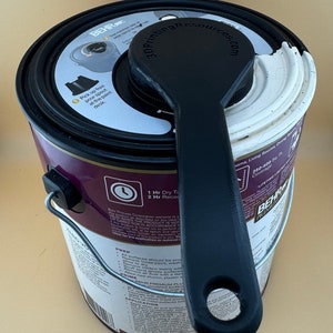 May include: A black paint can with a white lid and a black pour spout attached. The can is labeled "BEHR" and has a 1-hour dry time and a 2-hour recoat time. The can is sitting on a white surface.