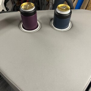 Puede incluir: Una mesa gris curvada con dos portavasos integrados. Cada soporte contiene un enfriador de latas, uno morado y otro azul marino, cada uno con una lata. La mesa tiene un borde cosido.