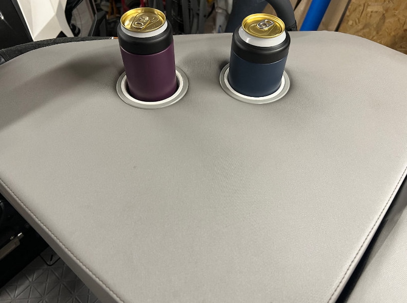 Puede incluir: Una mesa gris curvada con dos portavasos integrados. Cada soporte contiene un enfriador de latas, uno morado y otro azul marino, cada uno con una lata. La mesa tiene un borde cosido.