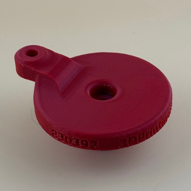May include: A red 3D printed plastic object with a hole in the centre and a small handle on the side. The object has the text "3D Printed" and "B30392" printed on the bottom.