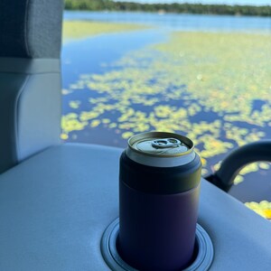 Puede incluir: Un enfriador de latas morado con una tapa negra sostiene una lata plateada. El enfriador est&aacute; en un portavasos gris sobre una superficie gris claro. El fondo muestra un lago con nen&uacute;fares y un cielo azul.