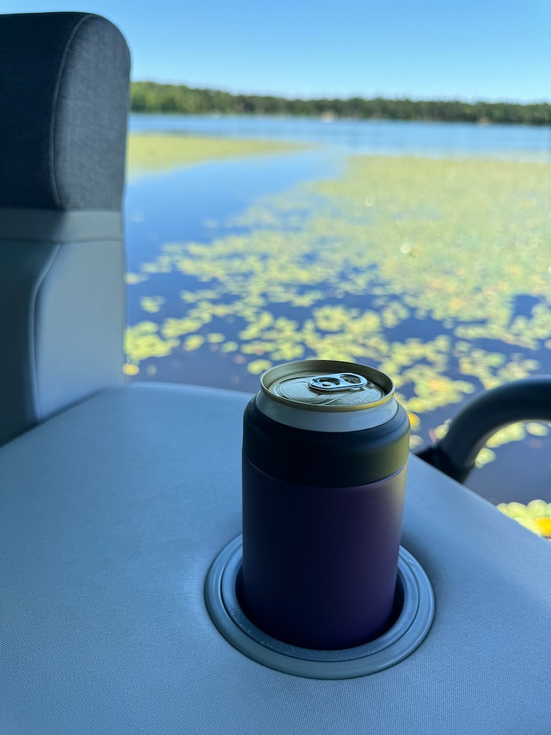 Puede incluir: Un enfriador de latas morado con una tapa negra sostiene una lata plateada. El enfriador est&aacute; en un portavasos gris sobre una superficie gris claro. El fondo muestra un lago con nen&uacute;fares y un cielo azul.
