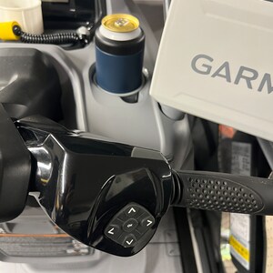 Puede incluir: Primer plano del manillar de una moto de nieve con un panel de control negro y un dispositivo Garmin. Un enfriador de latas azul y blanco contiene una bebida. El manillar tiene un agarre texturizado.