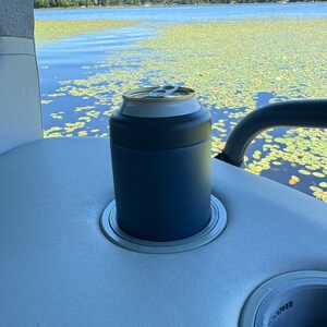 Puede incluir: Un enfriador de latas azul marino con un borde plateado, que sostiene una lata plateada y dorada. El enfriador est&aacute; colocado en el portavasos de un barco, con un lago y nen&uacute;fares en el fondo.