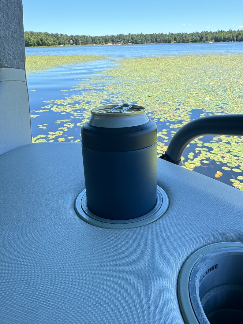 Puede incluir: Un enfriador de latas azul marino con un borde plateado, que sostiene una lata plateada y dorada. El enfriador est&aacute; colocado en el portavasos de un barco, con un lago y nen&uacute;fares en el fondo.