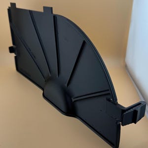 May include: A black, fan-shaped plastic component with a curved edge and several angled ridges. The piece has a semi-circular base and a rectangular section with tabs. The object is likely a part of a larger assembly.